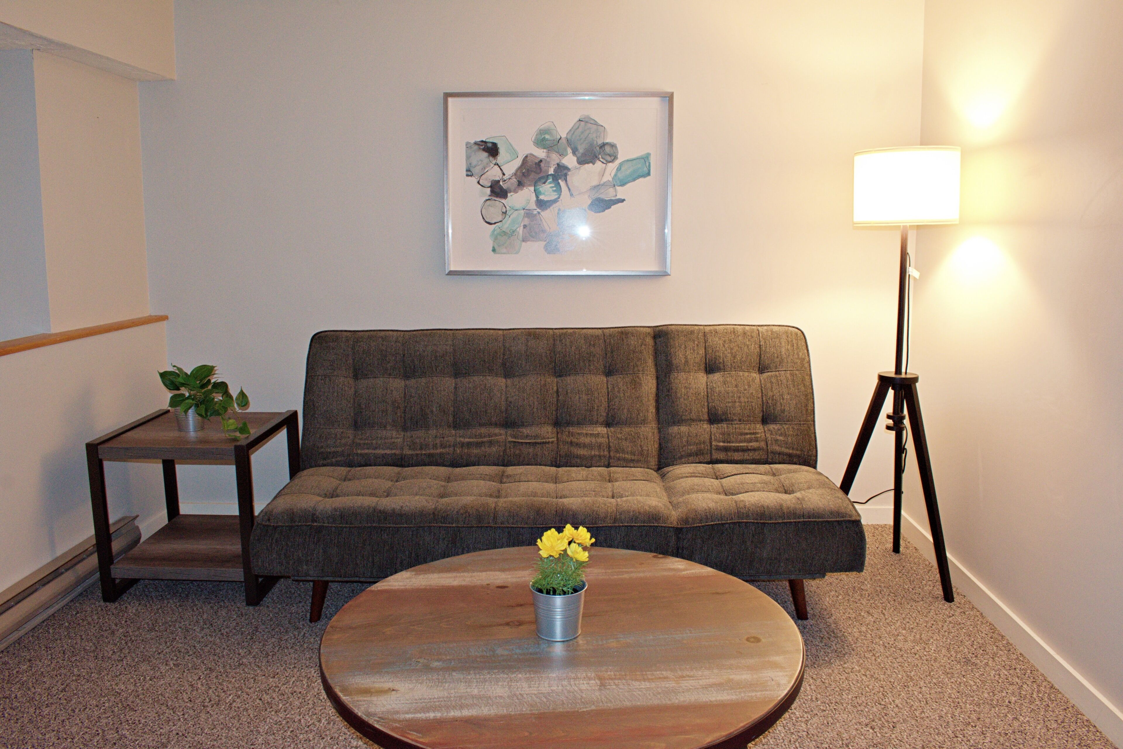 A living room with a brown sofa, a round coffee table with a potted plant, a small side table with a plant, a framed abstract artwork on the wall, and a floor lamp.