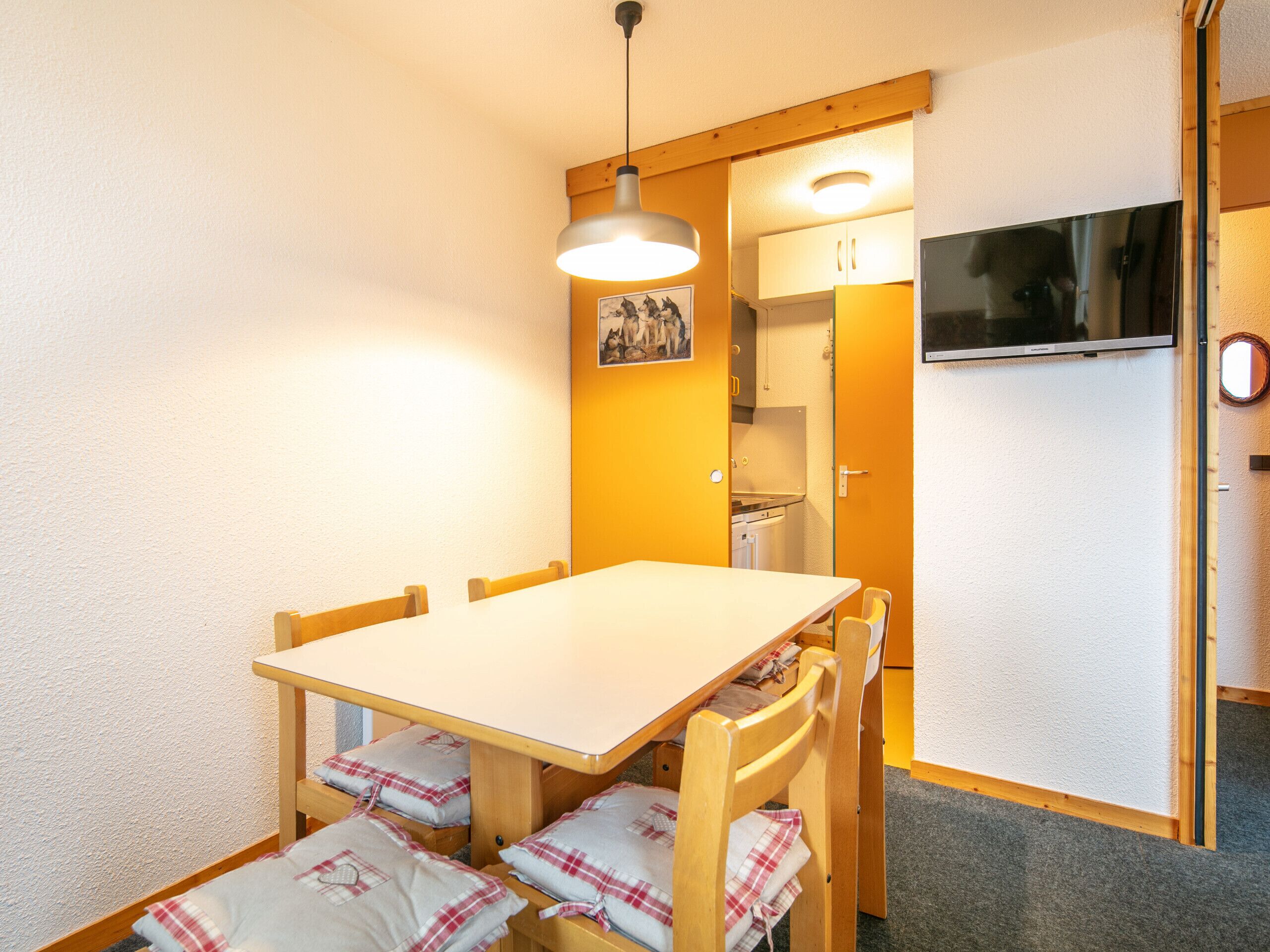 Habitación pequeña con una mesa de comedor de madera, cuatro sillas, una lámpara colgante, una zona de cocina y un televisor montado.