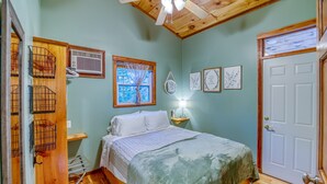 Une chambre à coucher avec un lit, un plancher en bois, un ventilateur de plafond, une fenêtre avec un rideau et une porte.