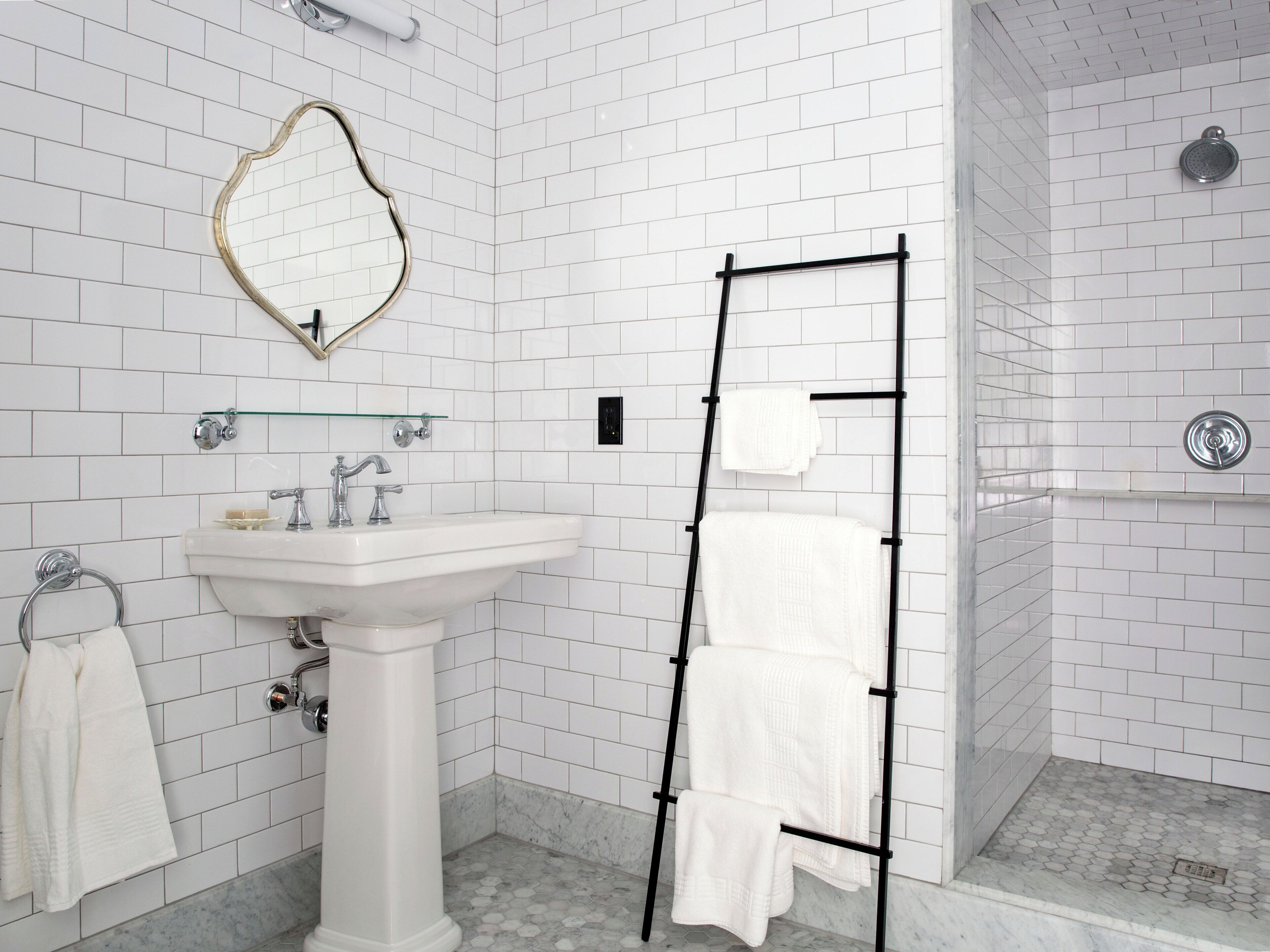 A bathroom with white tiled walls, a pedestal sink, a glass shelf, a towel rack, and a shower area.