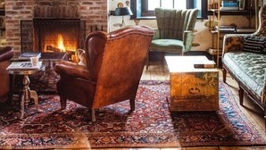 Un salon chaleureux avec une cheminée en briques, un fauteuil en cuir, un tapis à motifs et une bibliothèque remplie de livres.