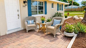 Une maison dont la porte porte le numéro 516, deux fauteuils en osier avec des coussins, une petite table et un jardin avec des plantes et des rochers.