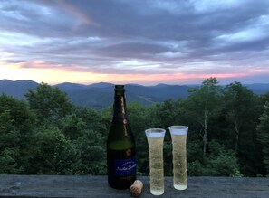 A bottle of beer and two glasses on a wooden surface with a scenic view of mountains and trees in the background.