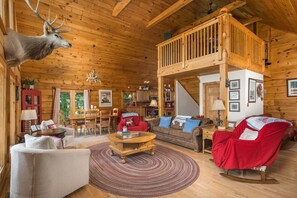 A log cabin interior with a dining area, a living room with a fireplace, and a loft space.