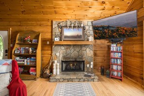 A cozy cabin interior with a stone fireplace, wooden bookshelves, and a large window with a scenic view.