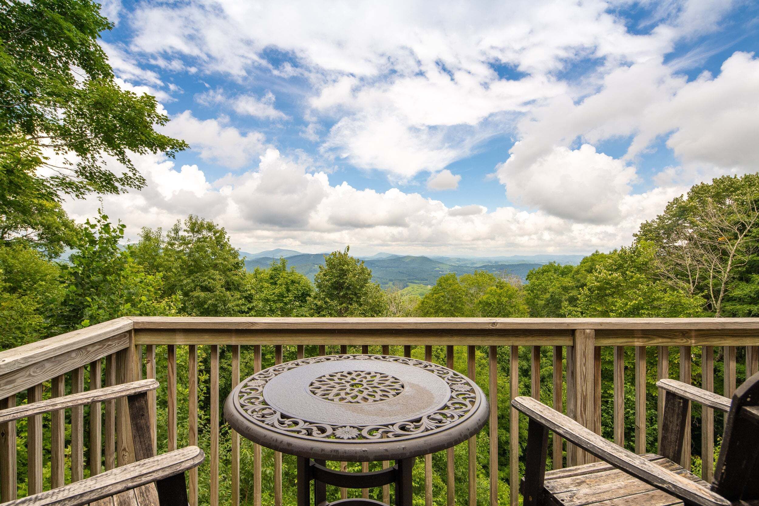 A wooden deck with a round table and chairs, overlooking a mountainous landscape with trees and a partly cloudy sky.