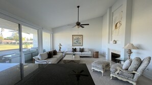 A spacious living room with a large window, a ceiling fan, a fireplace, and a dining area. - Golf Course Condo. X-Large 2 Bedroom (Rancho Mirage)