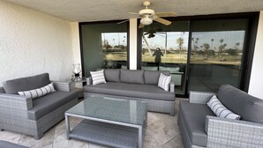 A covered patio area with a grey sectional sofa, a glass coffee table, and a ceiling fan. - Golf Course Condo. X-Large 2 Bedroom (Rancho Mirage)