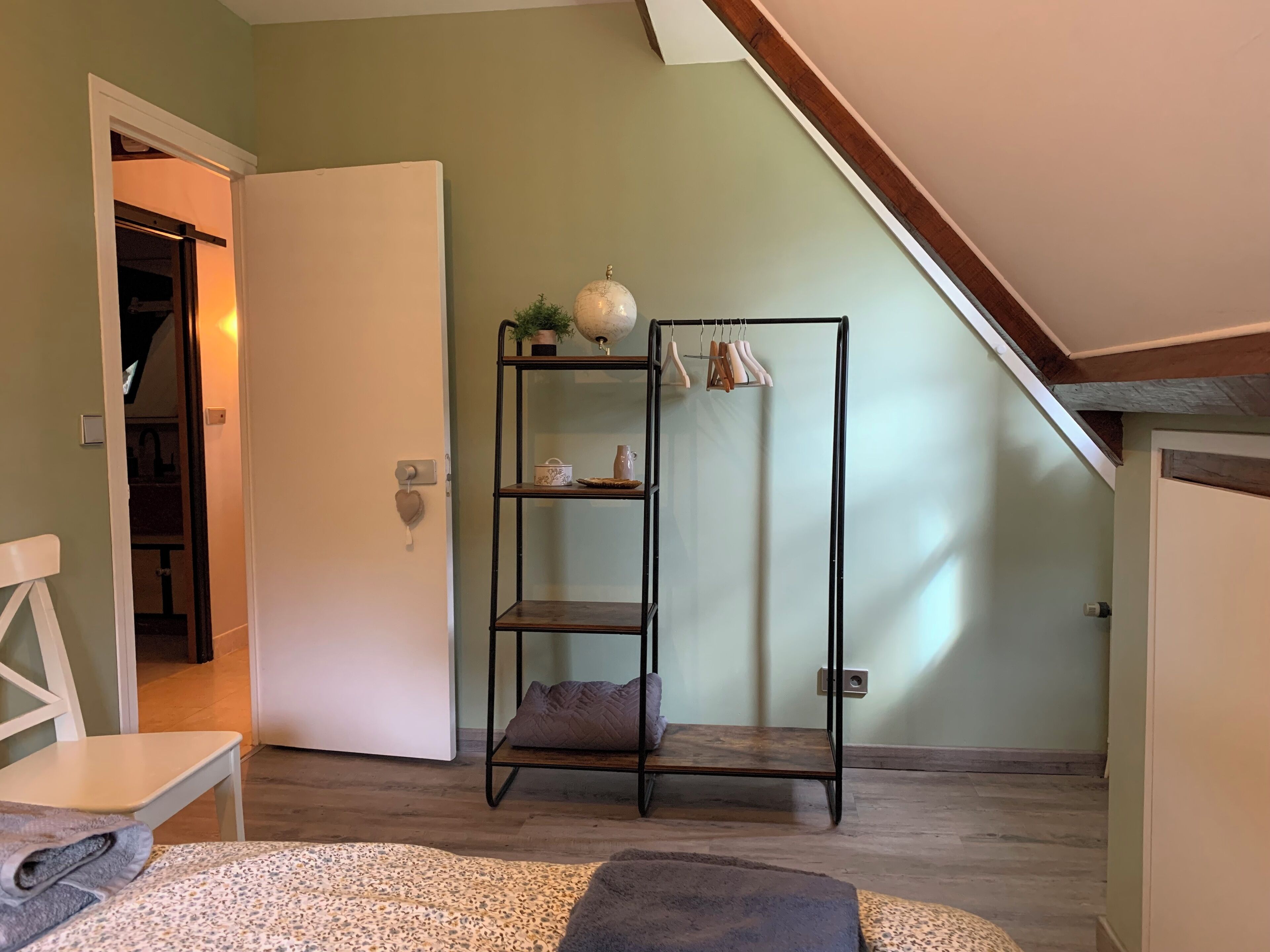Ein Zimmer mit einem Bett, einer weißen Tür, einem hohen Regal mit Gegenständen und einer geneigten Decke.
