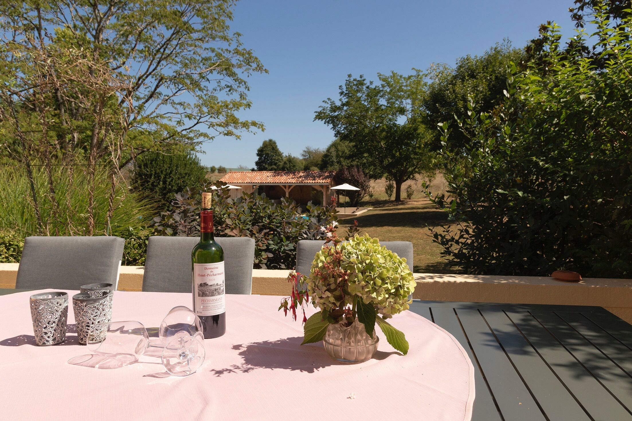 Une table à manger installée à l’extérieur, avec une bouteille de vin, des verres et un vase de fleurs.