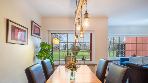 Un espace repas comprenant une table en bois, des chaises, des luminaires suspendus et une vue dégagée sur l’extérieur grâce à une grande fenêtre.