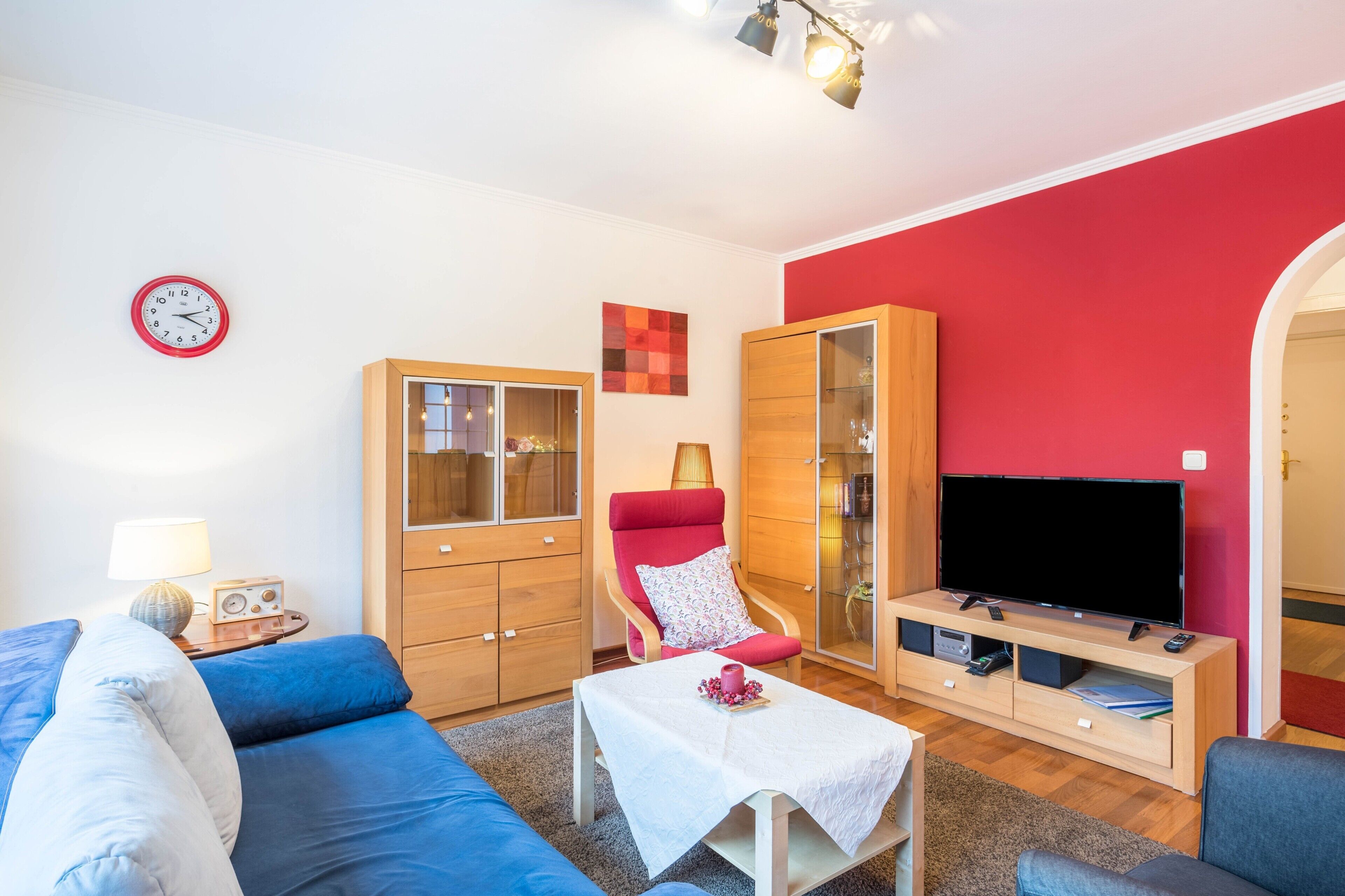 Ein Wohnzimmer mit einer Couch, einem roten Sessel, einem Fernseher, einem Schrank und einer Wanduhr.