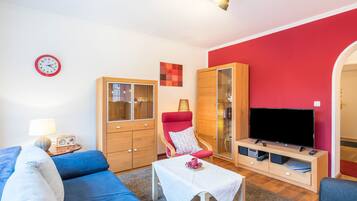 Ein Wohnzimmer mit einer Couch, einem roten Sessel, einem Fernseher, einem Schrank und einer Wanduhr.