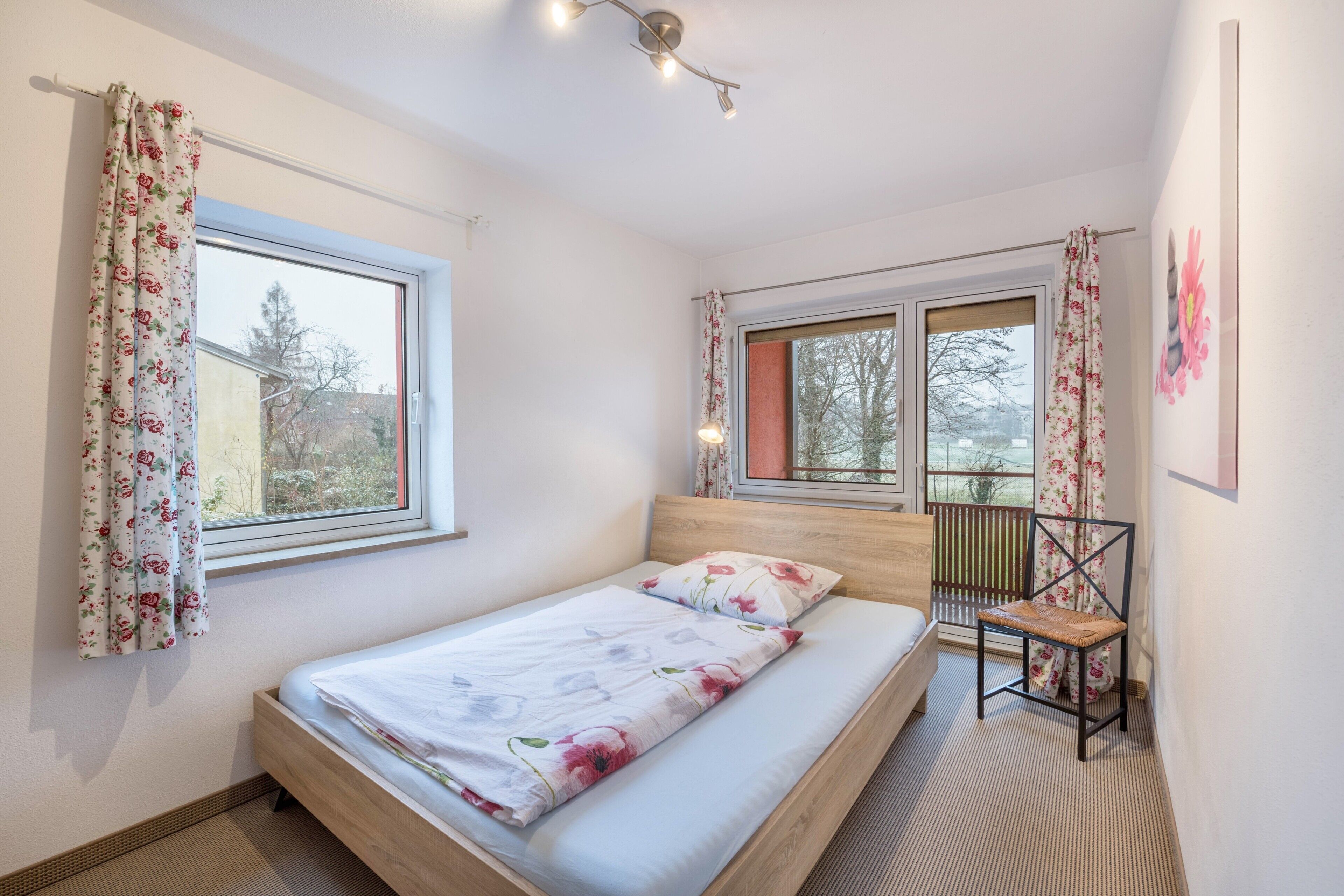 Ein Schlafzimmer mit einem Bett, zwei mit Vorhängen versehenen Fenstern, einem Sessel und einer Deckenleuchte.
