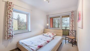 Ein Schlafzimmer mit einem Bett, zwei mit Vorhängen versehenen Fenstern, einem Sessel und einer Deckenleuchte.