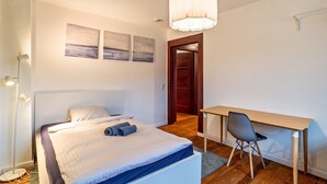 Une chambre à coucher avec un lit, un bureau et une chaise. Il y a des tableaux au mur, une lampe sur pied et une porte qui donne sur une autre pièce.