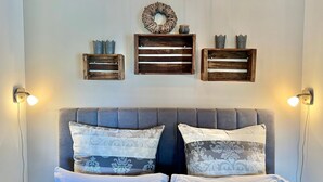 A bedroom with a grey upholstered headboard, two patterned pillows, and wall-mounted wooden shelves with decorative items. - fewo1846 - UferNest (App. 603) / Studio apartment with balcony on the 1st floor - (Harrislee)
