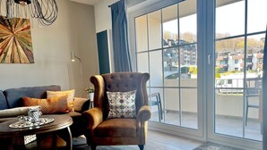 A modern living room with a large window, a brown armchair, a sofa, and a coffee table. - fewo1846 - UferNest (App. 603) / Studio apartment with balcony on the 1st floor - (Harrislee)