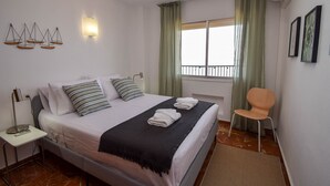 Ein ordentlich bezogenes Bett mit Kissen und Decke, ein Nachttisch mit Lampe, ein Stuhl und ein Fenster mit Vorhängen.