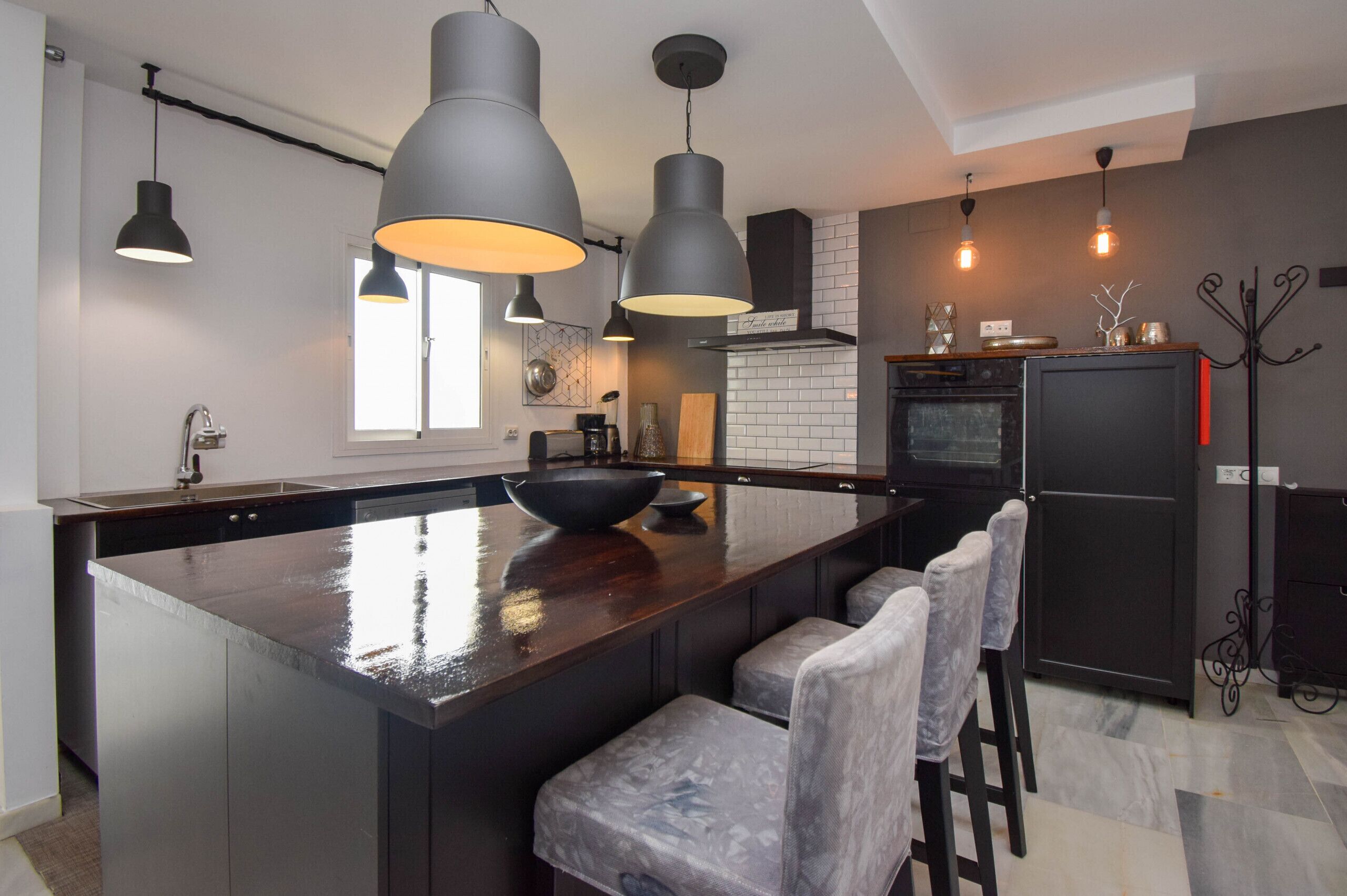 Una cocina moderna con una isla grande, taburetes de bar, iluminación colgante y un refrigerador negro.