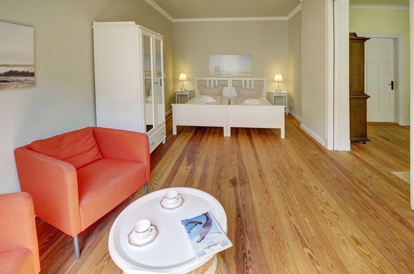 Habitación de hotel con un sillón rojo, una cama blanca con dos mesitas de noche, un armario blanco y suelo de madera.