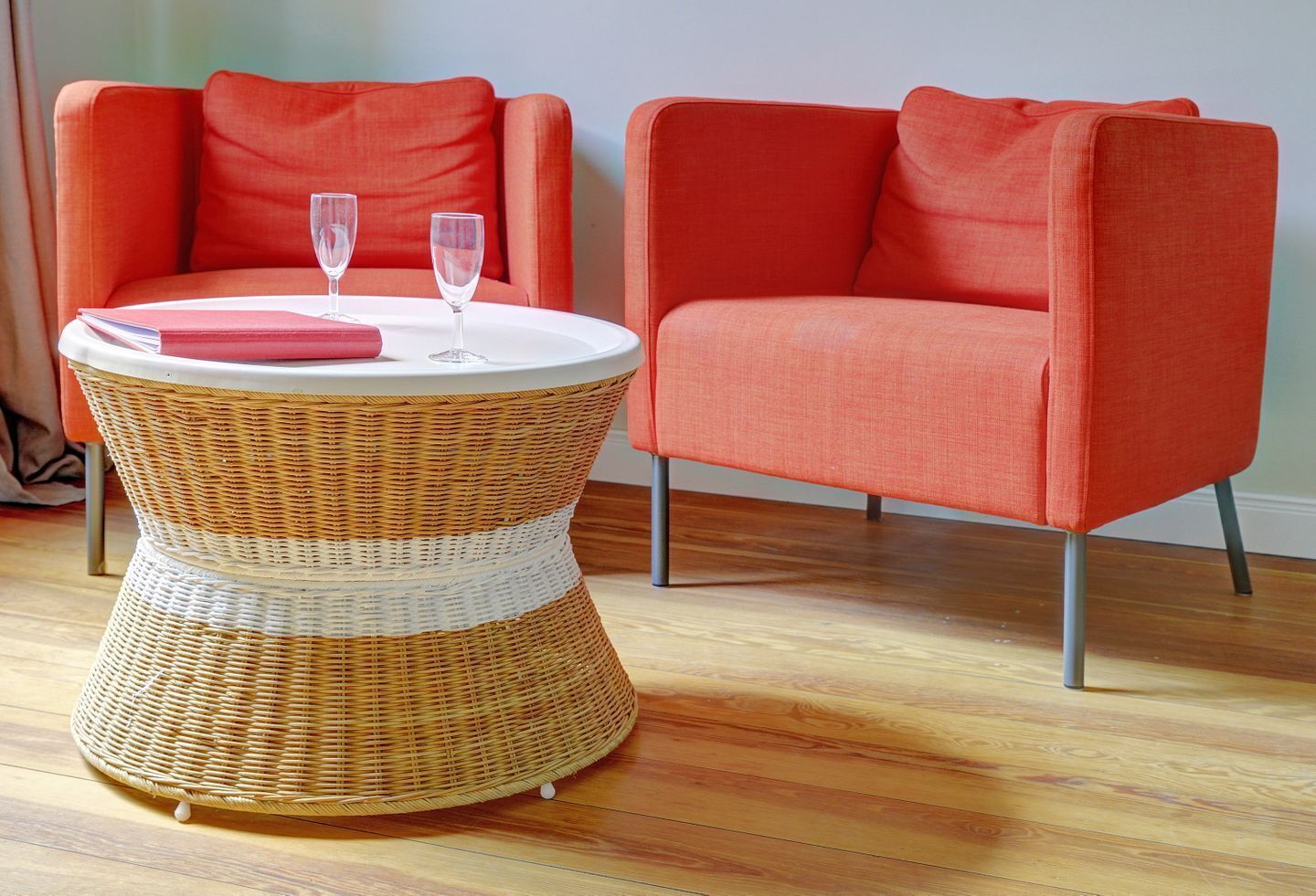 Una mesita de mimbre con dos vasos y una libreta rosa, acompañada por dos sillones naranjas.