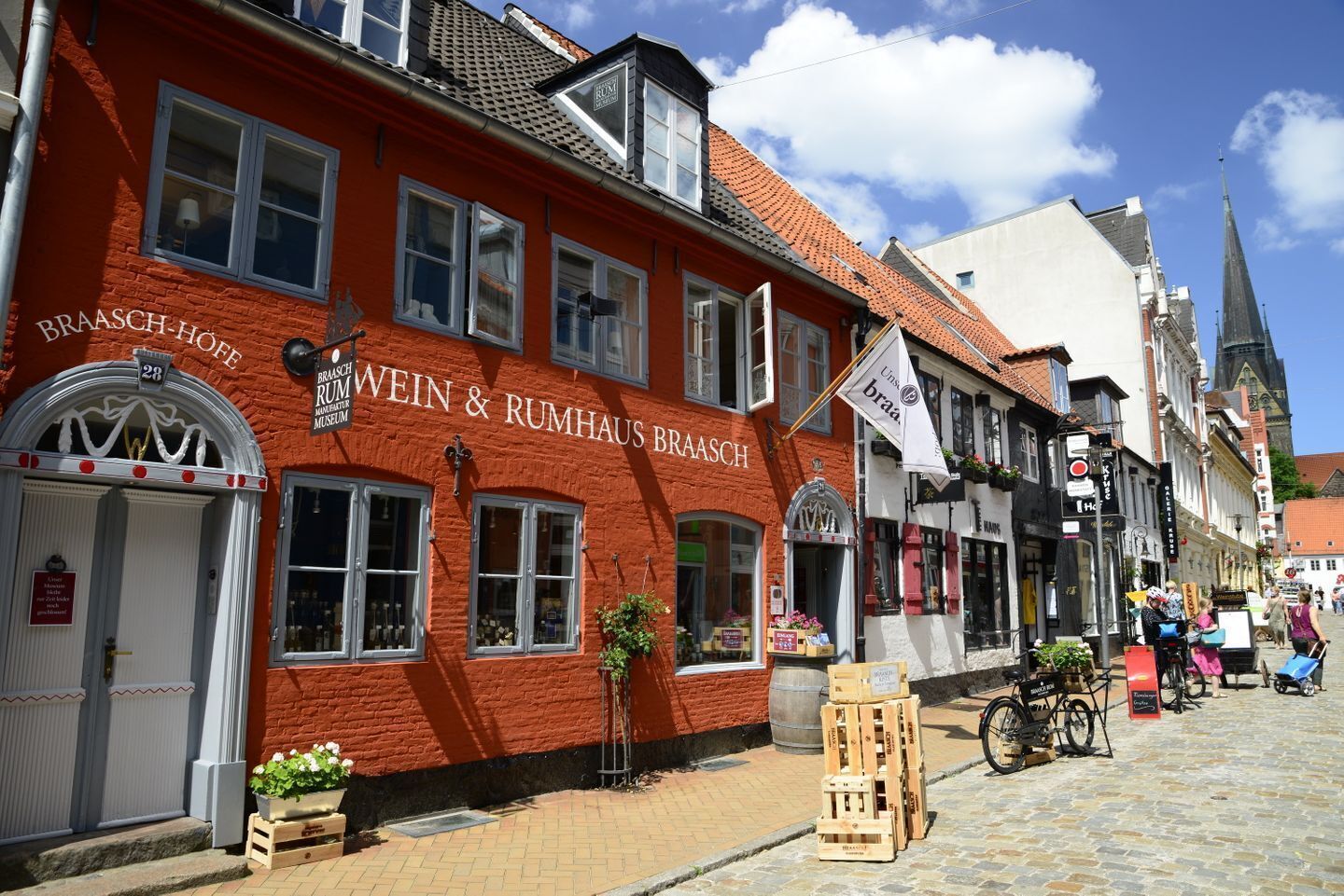 Un bâtiment rouge portant l’enseigne « BRAASCH-HOF WEIN & RUMHAUS BRAASCH » et un drapeau blanc avec le texte « Unss braa ».