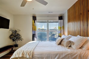 A bedroom with a bed, a potted plant, a ceiling fan, and a view of buildings through the window.