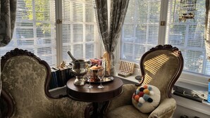 Una habitación con un ventanal grande, una mesa con tetera, vasos y libros, y una silla con un cojín colorido.