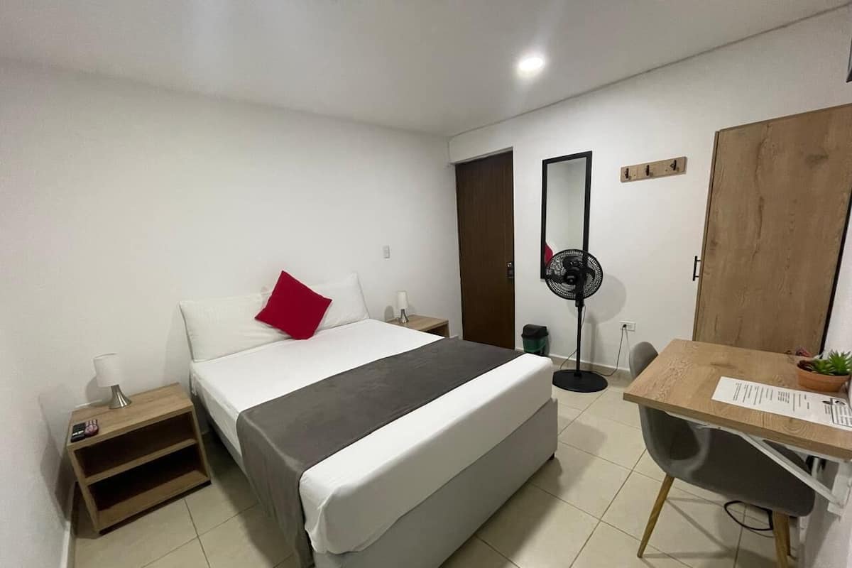 A hotel room with a bed, bedside table, desk, chair, and a standing fan.