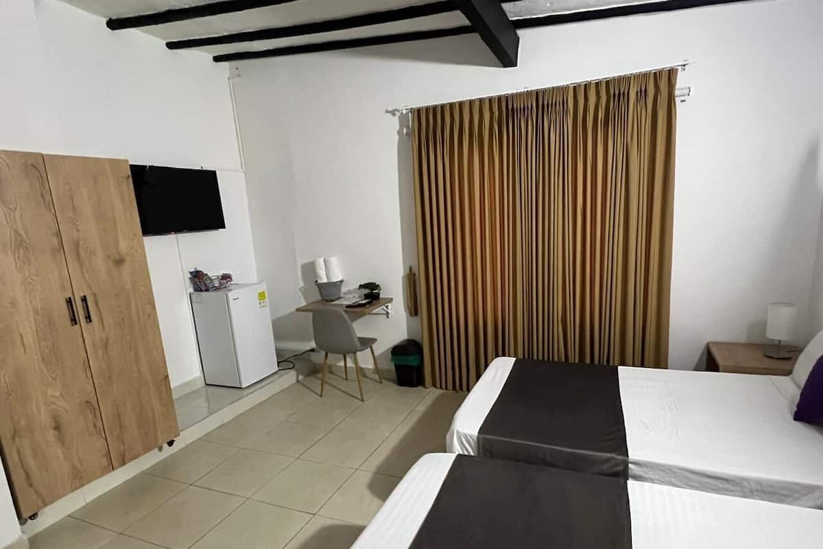 A hotel room with two beds, a wooden wardrobe, a desk with a chair, a small fridge, and a television mounted on the wall.