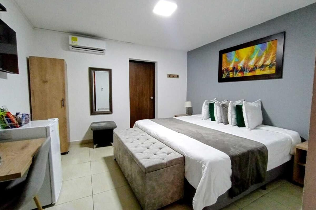 A hotel room with a bed, a desk, a chair, a small stool, a painting on the wall, and an air conditioning unit.