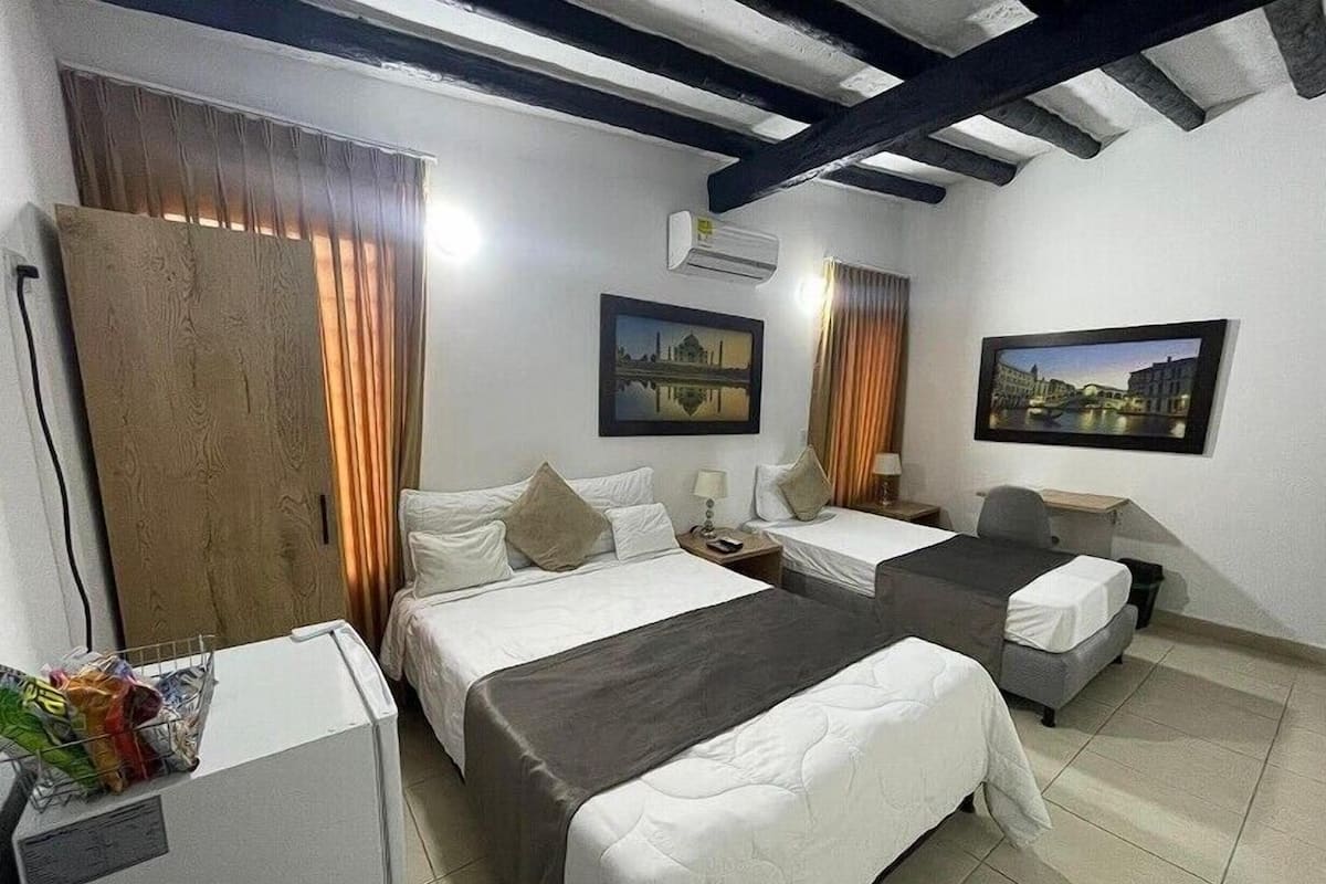 A hotel room with two beds, a desk, and a chair. There are curtains, a painting, and an air conditioning unit.