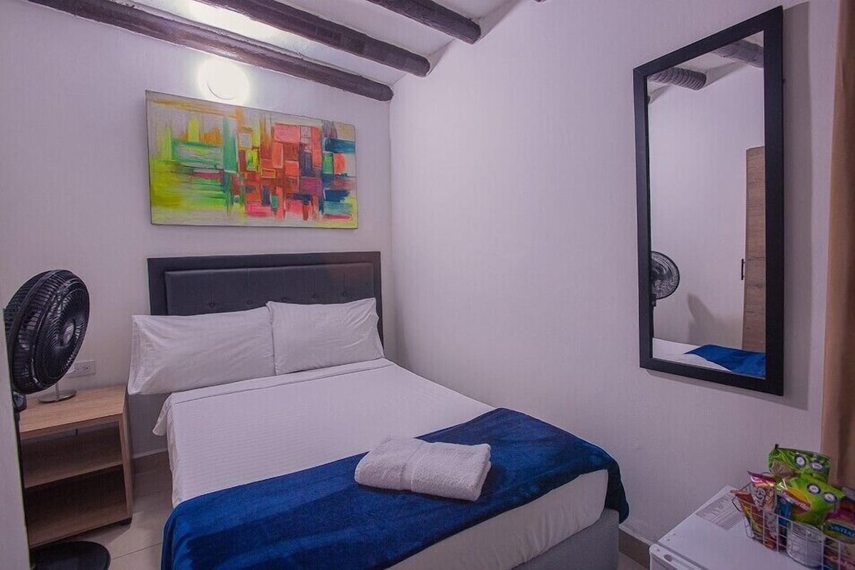 A hotel room with a bed, a fan, a mirror, and a colorful abstract painting on the wall.