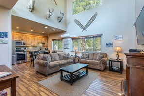 A spacious living area with a large sofa set, a coffee table, and a fireplace. There is a kitchen in the background with modern appliances. - Beautiful retreat on Hat Creek, Fire pits, Fishing and  15 min. from Lassen Park (Old Station)