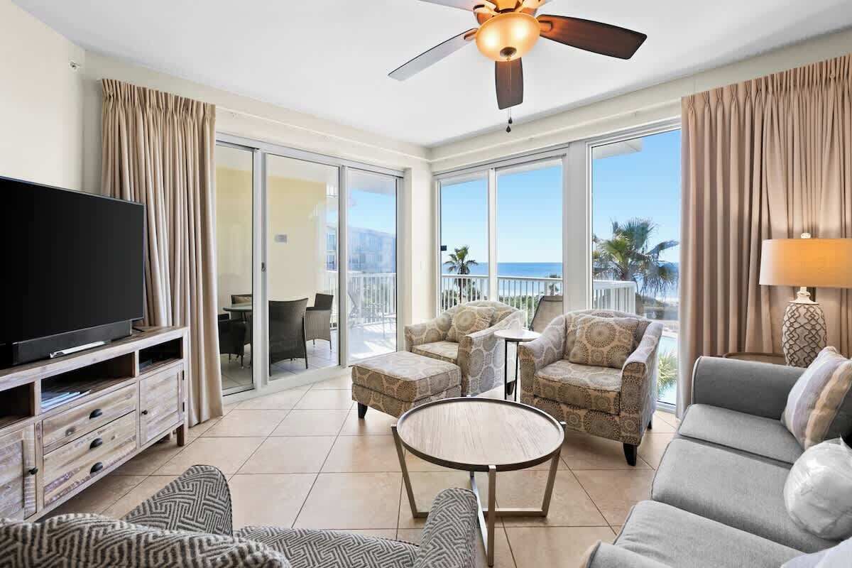 A spacious living room with a flat-screen TV, a ceiling fan, and a balcony with a view of the ocean.