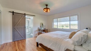A bedroom with a wooden floor, a bed with white bedding, a desk with a lamp, a ceiling fan, and a window with a view of trees.