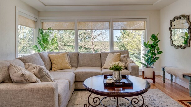 A living room with a sectional sofa, a round coffee table, and large windows with blinds.