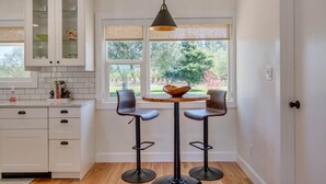 A kitchen with white cabinets, a round table with two bar stools, a hanging pendant light, and a window with a view of trees.