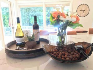 Una encimera de cocina con una botella de vino, una planta pequeña en maceta, un jarrón con flores y un cuenco de frutos secos.