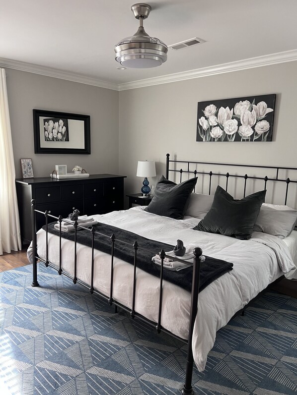 A neatly arranged bedroom with a bed, bedside tables, a nightstand, a ceiling fan, and artwork on the walls.