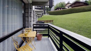 A balcony with yellow chairs and a table, overlooking a well-maintained lawn and traditional houses in the background.