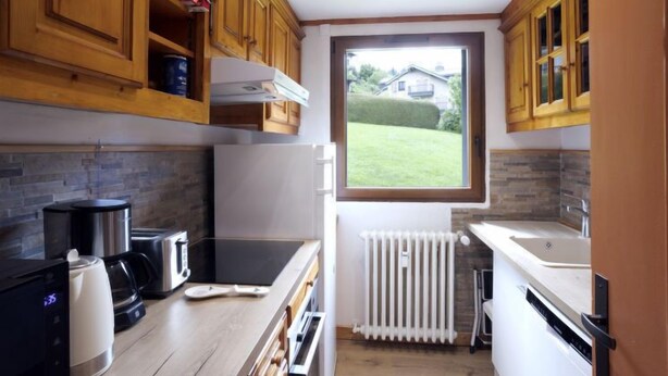 A compact kitchen with wooden cabinets, a built-in oven, a refrigerator, and a window overlooking a garden.