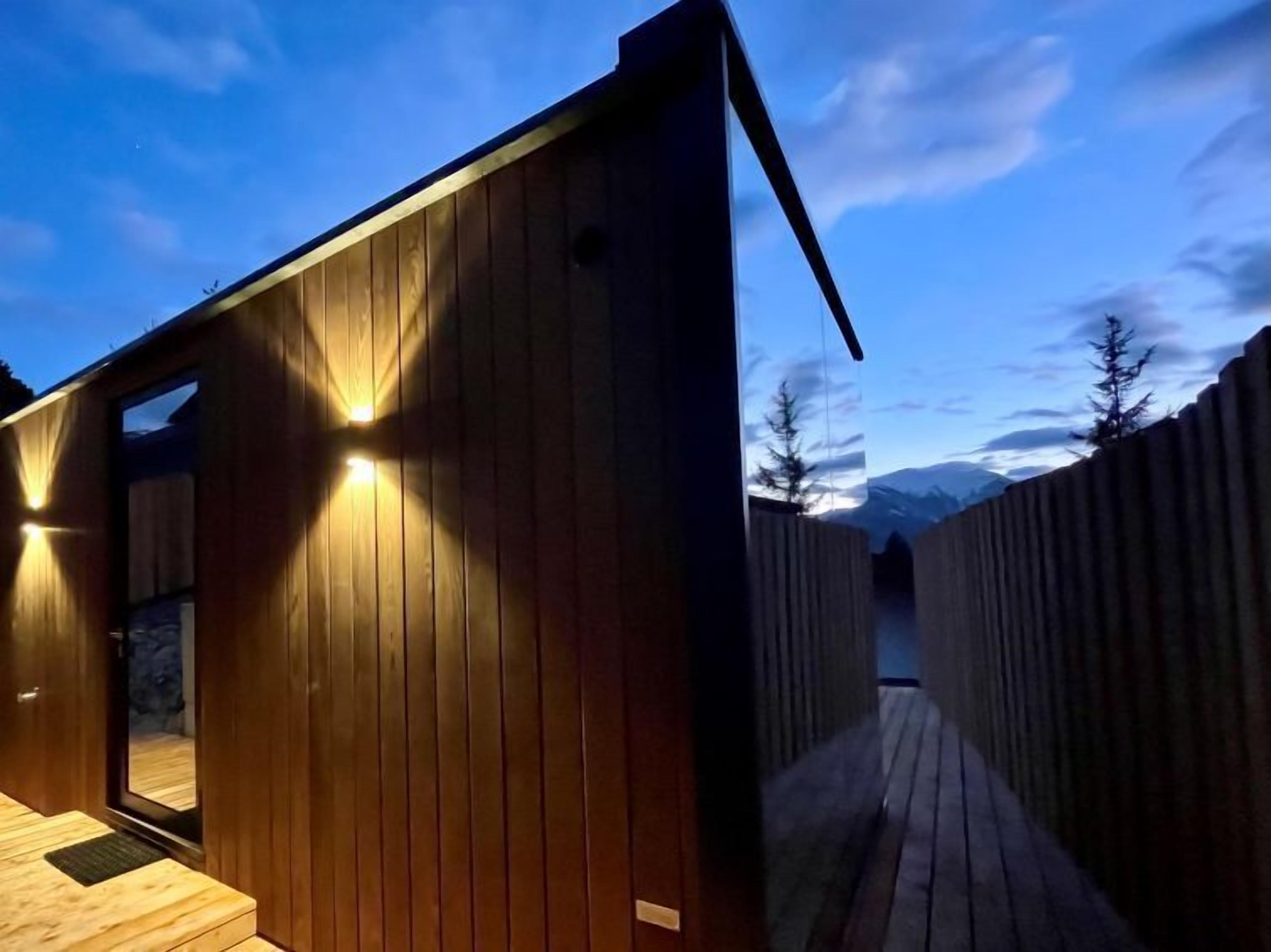 Una cabaña de madera moderna con terraza de madera y vista a montañas al fondo.