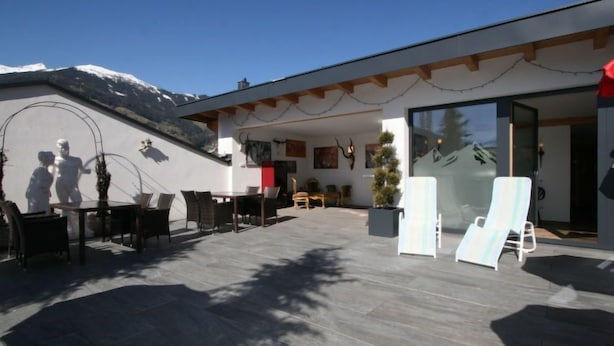 Zona salotto esterna moderna con vista sulle montagne, che include un tavolo da pranzo, sedie, un piccolo abete di Natale e un ingresso con porta in vetro.