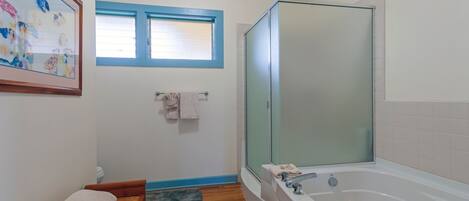 Un baño con bañera, ducha envuelta en vidrio, ventana con persianas y un cuadro en la pared.