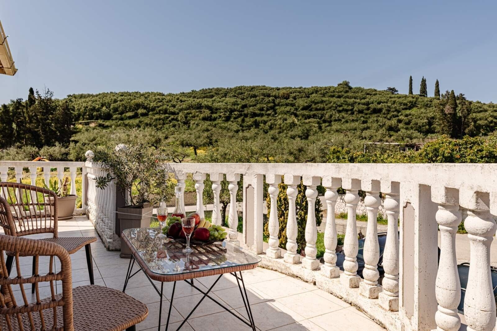 A terrace with wicker chairs, a glass table with fruit and wine glasses, and a white balustrade overlooking a lush hillside.