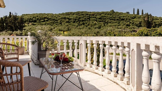 Une terrasse avec des chaises en osier, une table en verre ornée de fruits et de verres à vin, et une balustrade blanche donnant sur une colline verdoyante.