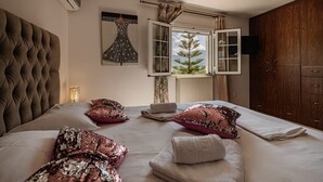 Une chambre à coucher avec une tête de lit capitonnée, un grand lit agrémenté d’oreillers décoratifs, une fenêtre donnant sur un arbre et une armoire en bois.