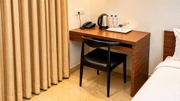 A hotel room with a wooden desk, a black chair, a bed with white bedding, a kettle, two water bottles, and a phone on the desk.